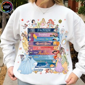 Puede incluir: Sudadera blanca con un diseño de cintas VHS apiladas, cada una con títulos de películas de Disney como "Frozen" y "Aladdin". Ilustraciones de princesas Disney. Cuello redondo y mangas largas.