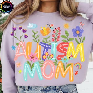 以下が含まれることがあります： 鮮やかな花柄の文字で「AUTISM MOM」と書かれた、薄紫色のスウェットシャツ。花、蝶、虹、パズルのピースがデザインされています。