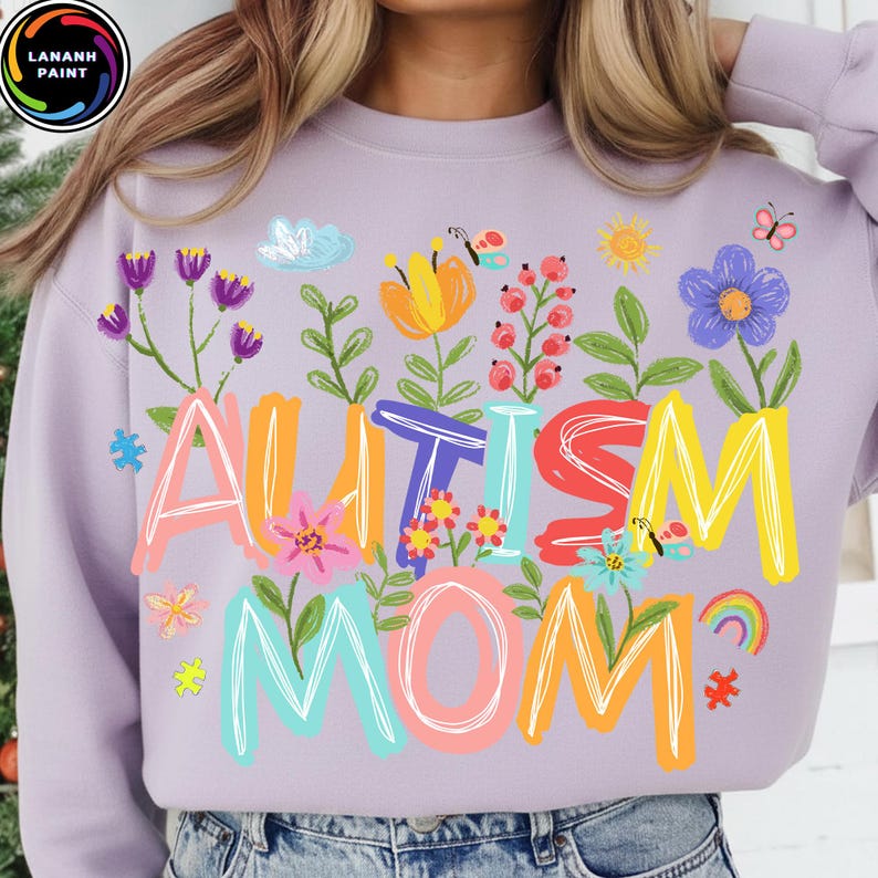 以下が含まれることがあります： 鮮やかな花柄の文字で「AUTISM MOM」と書かれた、薄紫色のスウェットシャツ。花、蝶、虹、パズルのピースがデザインされています。