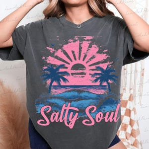 Pu&ograve; includere: Una maglietta grigia scura con una grafica di tramonto rosa e blu e palme. Il testo "Salty Soul" &egrave; stampato in rosa sotto la grafica.