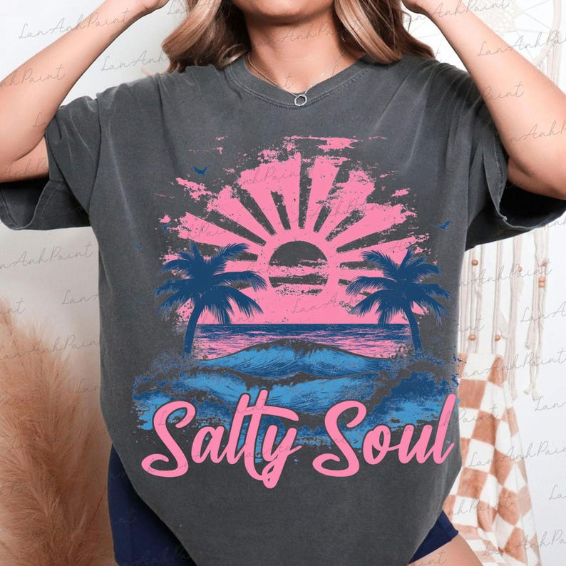 Pu&ograve; includere: Una maglietta grigia scura con una grafica di tramonto rosa e blu e palme. Il testo "Salty Soul" &egrave; stampato in rosa sotto la grafica.