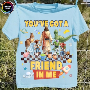 Può includere: Una t-shirt azzurra con una stampa grafica di vari personaggi dei cartoni animati e una figura centrale in tunica che si allontana, con il testo "YOU'VE GOT A FRIEND IN ME" in lettere gialle. Il design include pianeti, razzi e stelle.