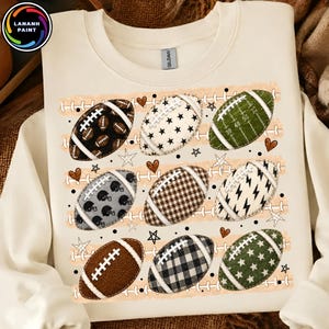 May include: A cream-colored crewneck sweatshirt featuring a graphic print of nine American footballs arranged in three rows. The footballs display various patterns including plaid, stars, lightning bolts, and solid colors, interspersed with small hearts and stars.