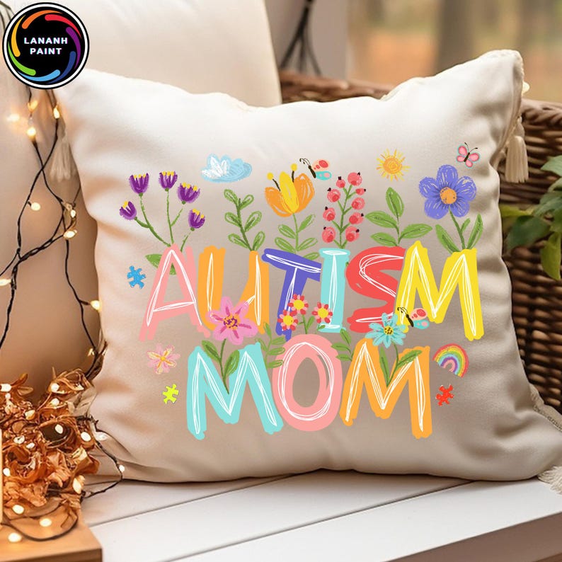 以下が含まれることがあります： カラフルな文字で「AUTISM MOM」と書かれたベージュ色のクッション。花や蝶のイラストで囲まれています。白い表面に置かれています。