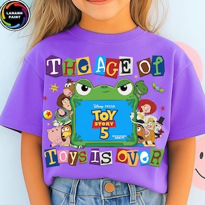 Puede incluir: Una camiseta morada de manga corta con un gr&aacute;fico de una rana verde que sostiene un cartel azul que dice Disney Pixar Toy Story 5. El texto The Age Of Toys Is Over rodea a la rana, junto con varias ilustraciones de personajes de Toy Story.