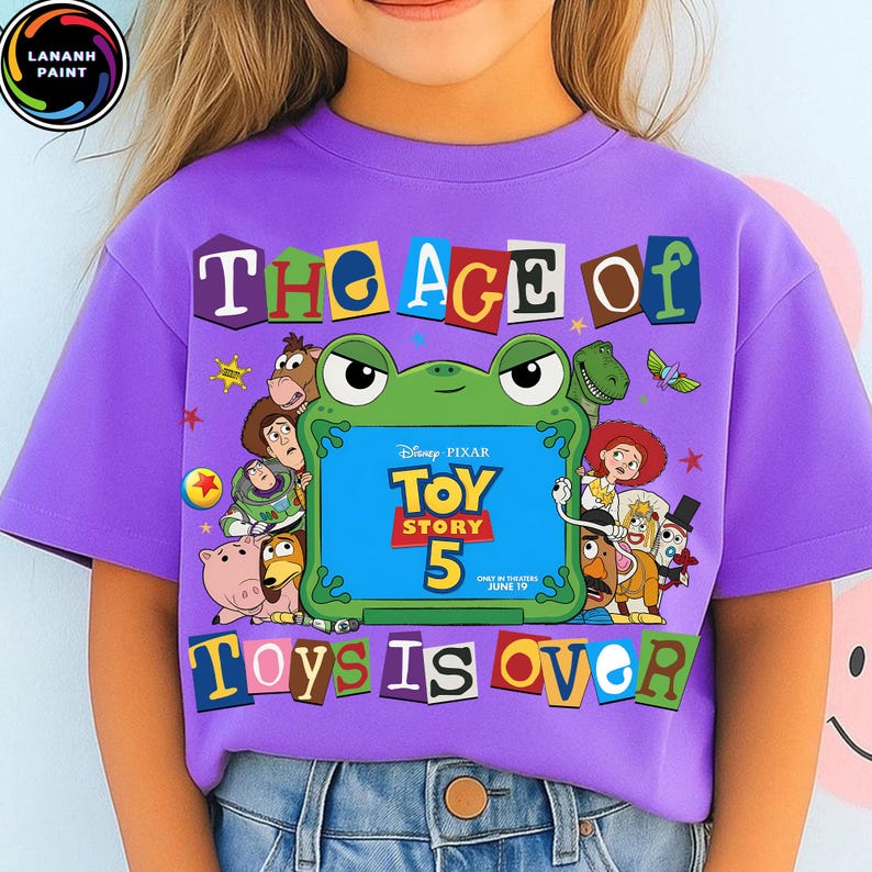 Puede incluir: Una camiseta morada de manga corta con un gr&aacute;fico de una rana verde que sostiene un cartel azul que dice Disney Pixar Toy Story 5. El texto The Age Of Toys Is Over rodea a la rana, junto con varias ilustraciones de personajes de Toy Story.
