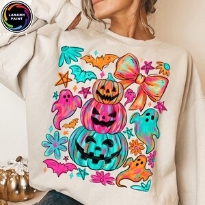 May include: Beige sweatshirt with a colorful Halloween-themed graphic. The design features stacked jack-o'-lanterns, ghosts, bats, stars, and a large bow in vibrant shades of pink, teal, orange, and yellow. The brand logo is in the upper left corner.