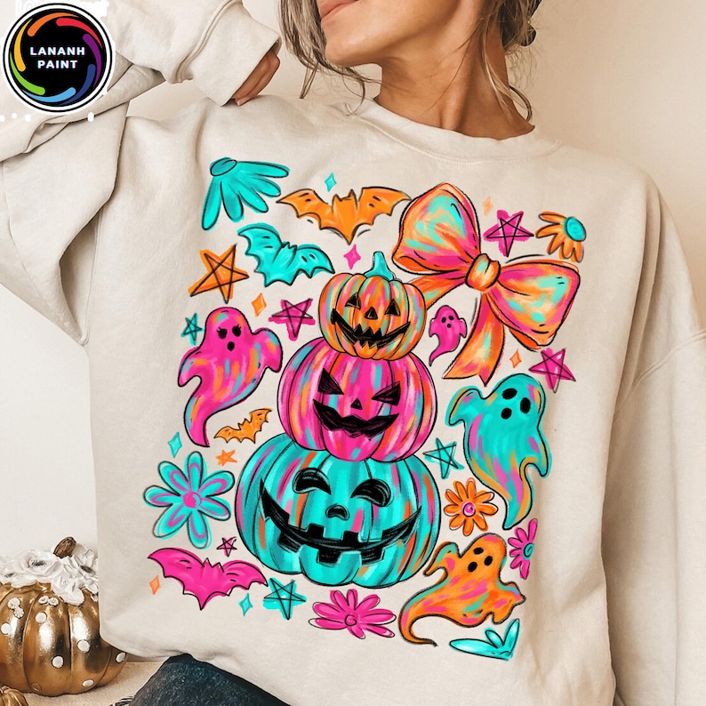 May include: Beige sweatshirt with a colorful Halloween-themed graphic. The design features stacked jack-o'-lanterns, ghosts, bats, stars, and a large bow in vibrant shades of pink, teal, orange, and yellow. The brand logo is in the upper left corner.
