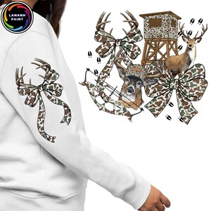 May include: White sweatshirt with a deer-themed design. The design includes a deer head, bow and arrow, camouflage bows, a deer stand, and a deer. The design is in shades of brown, green, and white.