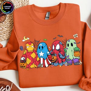 May include: An orange crewneck sweatshirt featuring a graphic print of four characters dressed as ghosts for Halloween. The characters resemble Iron Man, Captain America, Spider-Man, and a classic ghost, each holding a small orange pumpkin bucket.