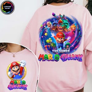 Puede incluir: Sudadera rosa claro con un gr&aacute;fico colorido de Mario y otros personajes del juego Super Mario Galaxy. El texto "THE SUPER MARIO GALAXY" se muestra en una fuente vibrante y multicolor.