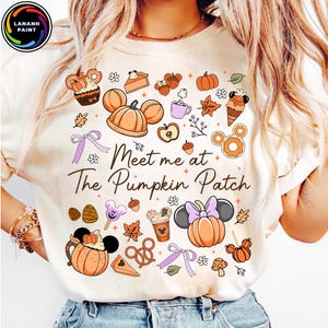 Może przedstawiać: Kremowa koszulka z kolorową grafiką w jesiennym stylu. Projekt zawiera dynie, uszy Myszki Miki, jesienne liście i napis "Meet me at The Pumpkin Patch". Koszulka jest ozdobiona różnymi produktami spożywczymi.