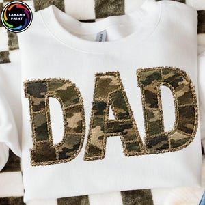 May include: White sweatshirt with the word "DAD" in large, textured, camouflage-patterned letters. The letters are outlined with a stitched, tan-colored border. The sweatshirt is folded on a checkered surface.