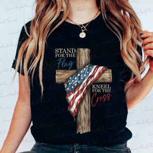 Puede incluir: Camiseta negra con una cruz de madera adornada con una bandera estadounidense. La camiseta presenta las frases "STAND FOR THE Flag" y "KNEEL FOR THE Cross" en texto azul y rojo.