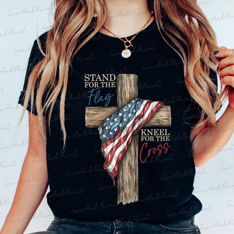 Puede incluir: Camiseta negra con una cruz de madera adornada con una bandera estadounidense. La camiseta presenta las frases "STAND FOR THE Flag" y "KNEEL FOR THE Cross" en texto azul y rojo.