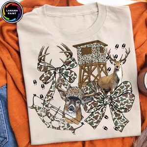 May include: Beige t-shirt featuring a hunting-themed design. The graphic includes a deer, a hunting blind, a bow and arrow, and decorative camouflage bows. The design incorporates brown, green, and white colors, with deer tracks scattered around the image.