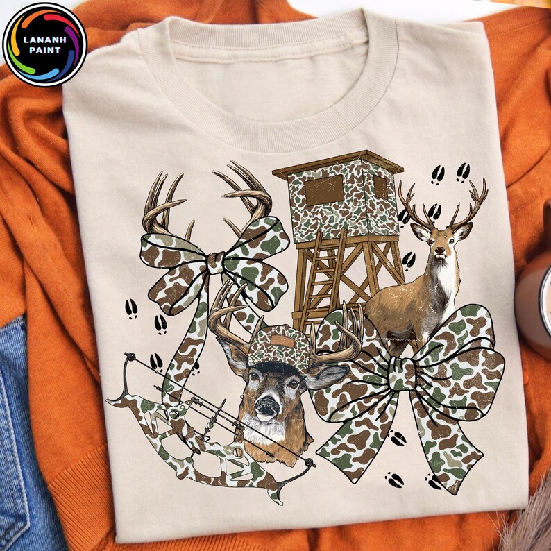May include: Beige t-shirt featuring a hunting-themed design. The graphic includes a deer, a hunting blind, a bow and arrow, and decorative camouflage bows. The design incorporates brown, green, and white colors, with deer tracks scattered around the image.