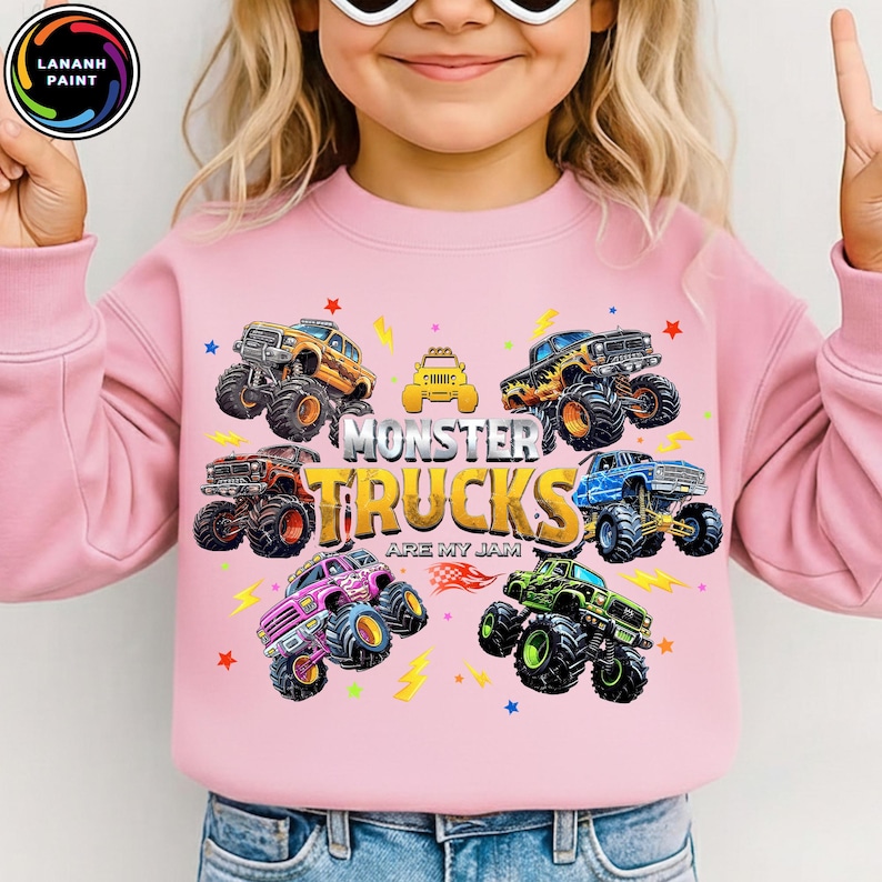 K&ouml;nnte beinhalten: Rosa Sweatshirt mit einem farbenfrohen Monster-Truck-Motiv. Das Design zeigt verschiedene Monster-Truck-Illustrationen, Sterne, Blitze und den Text "MONSTER TRUCKS ARE MY JAM."