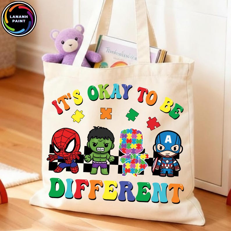 May include: Cream-colored tote bag with the text "IT'S OKAY TO BE DIFFERENT" in colorful letters. The bag features cartoon images of Spider-Man, Hulk, a puzzle piece figure, and Captain America. A purple teddy bear and books are inside.