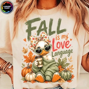 May include: A cream-colored t-shirt featuring a graphic of a duck wearing a floral bow, green sunglasses, and a green sweater, holding a coffee cup. The text reads FALL is my Love Language, surrounded by orange and green autumn leaves and pumpkins.