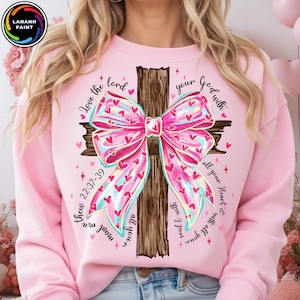 Könnte beinhalten: Hellrosa Sweatshirt mit einem Holzkreuz-Design und einer großen rosa Schleife, die mit roten Herzen verziert ist. Der Text lautet "Love the Lord your God with all your heart & with all your soul & with all your mind ma thew 22:37-39".