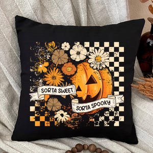 May include: Black square pillow with a Halloween design. The pillow features an orange jack-o'-lantern with the words "Sorta Sweet Sorta Spooky" on a banner. The design includes flowers and a checkered pattern.
