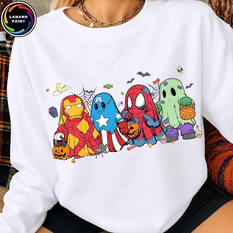 May include: A white crewneck sweatshirt featuring a graphic print of four characters dressed as ghosts for Halloween. The characters resemble Iron Man, Captain America, Spider-Man, and a classic ghost, each holding a small orange pumpkin bucket.