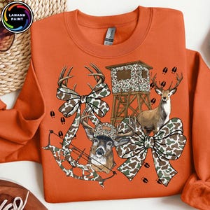 May include: Orange sweatshirt with a deer hunting theme. The design includes a deer, a hunting blind, camouflage bows, and a bow and arrow. The sweatshirt has the text "LANANH PAINT" in a circular logo in the upper left corner.
