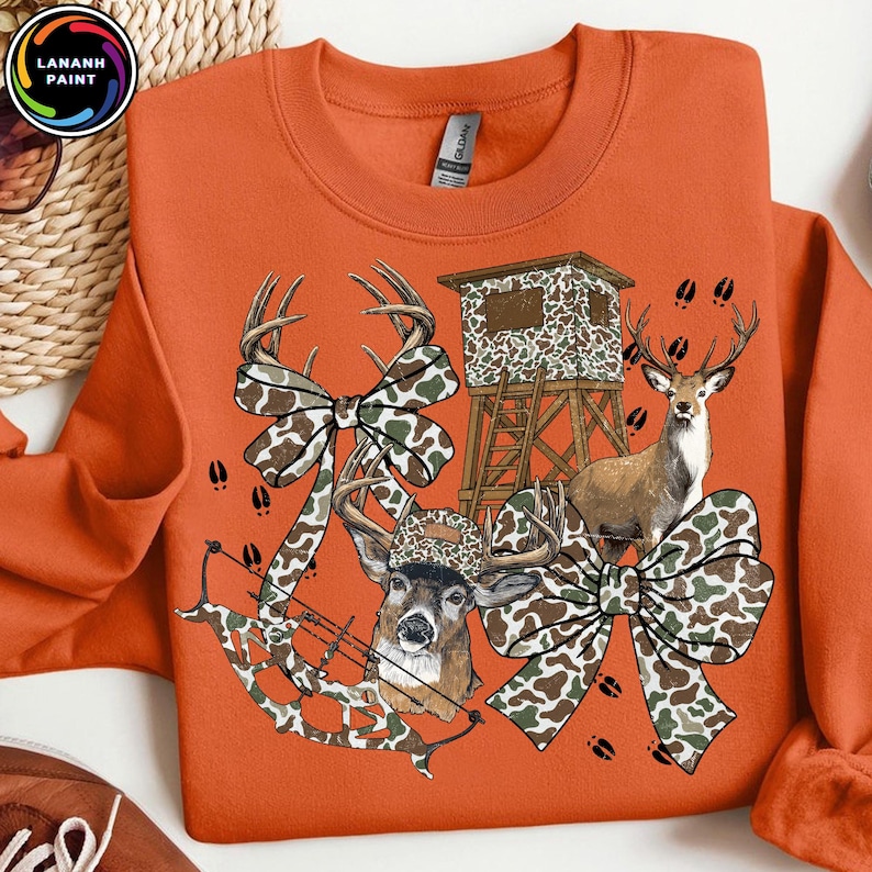 May include: Orange sweatshirt with a deer hunting theme. The design includes a deer, a hunting blind, camouflage bows, and a bow and arrow. The sweatshirt has the text "LANANH PAINT" in a circular logo in the upper left corner.