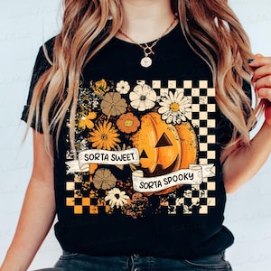 May include: Black t-shirt featuring a pumpkin with a smiling face, surrounded by flowers in shades of orange, yellow, and brown. The text "Sorta Sweet Sorta Spooky" is displayed on a banner. A checkered pattern is on the right side.