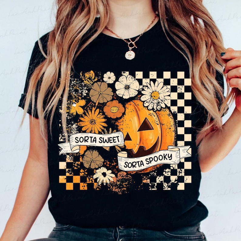 May include: Black t-shirt featuring a pumpkin with a smiling face, surrounded by flowers in shades of orange, yellow, and brown. The text "Sorta Sweet Sorta Spooky" is displayed on a banner. A checkered pattern is on the right side.