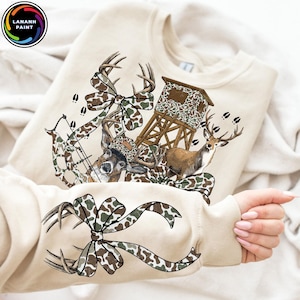 May include: A beige sweatshirt featuring a hunting-themed graphic. The design includes a deer, a bow and arrow, a hunting blind, and a camouflage bow. The text "LANANH PAINT" is in a circle in the upper left corner.