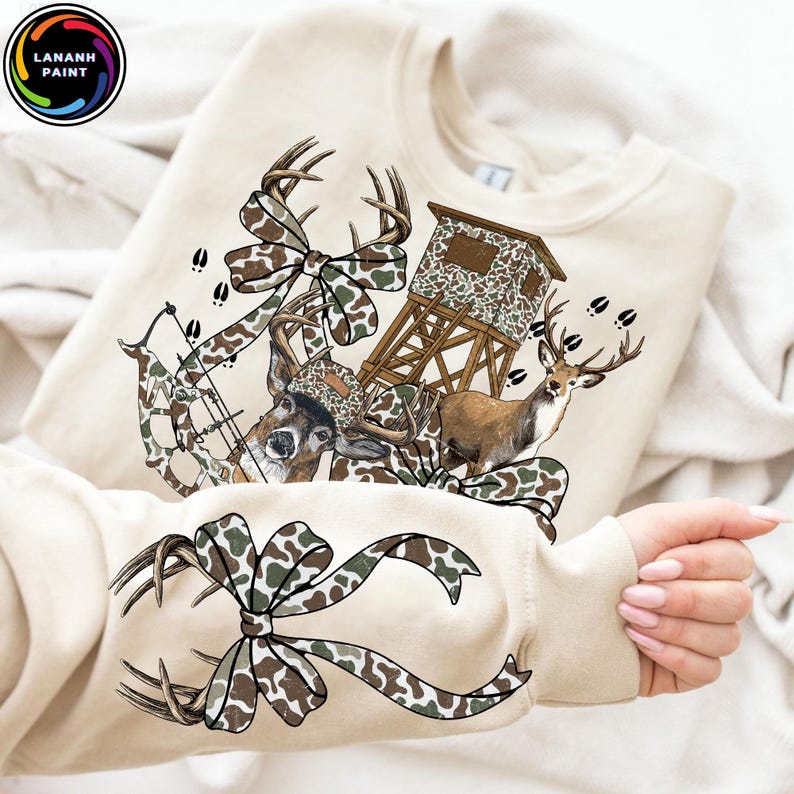 May include: A beige sweatshirt featuring a hunting-themed graphic. The design includes a deer, a bow and arrow, a hunting blind, and a camouflage bow. The text "LANANH PAINT" is in a circle in the upper left corner.