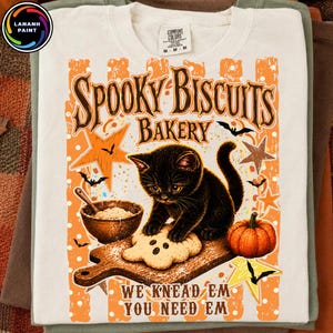 May include: A cream-colored t-shirt featuring a graphic of a black kitten on a wooden cutting board with a ghost-shaped cookie, a bowl of flour, and a pumpkin. The text reads "SPOOKY BISCUITS BAKERY" and "WE KNEAD EM YOU NEED EM" in brown.