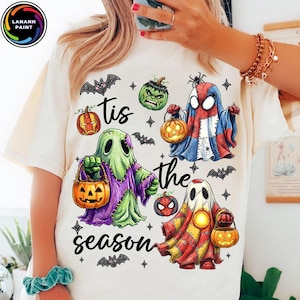 May include: A cream-colored t-shirt featuring a graphic print with the text 'tis the season' and illustrations of three ghosts dressed as superheroes, each holding a jack-o'-lantern, surrounded by small bats, stars, and pumpkin icons.