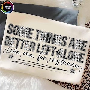 Puede incluir: Camiseta color crema con el texto "SOME THINGS ARE BETTER LEFT ALONE Like me, for instance." El texto presenta patrones de cuadros, leopardo y estrellas. La camiseta se muestra sobre una superficie blanca y suave.