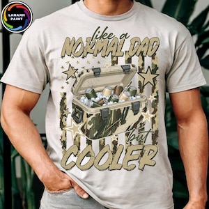May include: A light gray t-shirt featuring a graphic of a camouflage-patterned cooler filled with beverage cans and ice. The design includes the text "like a NORMAL DAD but COOLER" in a distressed font, surrounded by stars and vertical stripes.