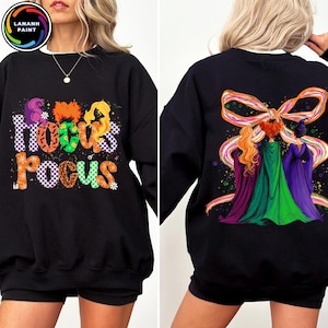 May include: Black sweatshirt featuring the text "Hocus Pocus" in colorful, patterned letters on the front. The back of the sweatshirt displays an illustration of three figures with long hair and capes, with a large ribbon design.