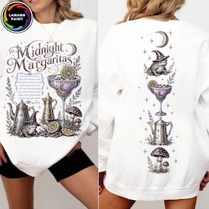 May include: A white long-sleeve sweatshirt featuring a graphic print with the text Midnight Margaritas. The design includes a frog wearing a witch hat, a margarita glass, a teapot, mushrooms, and a crescent moon in shades of purple and black.