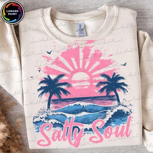 Pu&ograve; includere: Una felpa color crema con un design grafico rosa e blu di un tramonto su una spiaggia con palme e onde. Il testo "Salty Soul" &egrave; scritto in lettere rosa sotto il grafico.