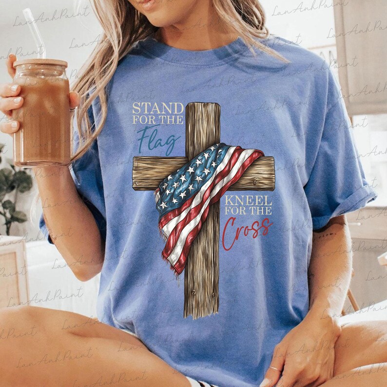 Puede incluir: Camiseta azul claro con una cruz de madera y una bandera estadounidense. El texto dice "STAND FOR THE Flag" y "KNEEL FOR THE Cross". La camiseta es de estilo casual.