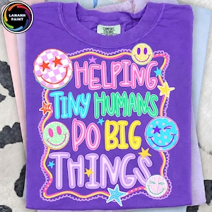 Op de afbeelding: Een paars t-shirt met een grafische print met de tekst HELPING TINY HUMANS DO BIG THINGS. Het ontwerp bevat kleurrijke smileys met ruitjes-, stippen- en streepjespatronen, samen met verschillende stervormen binnen een golvende rand.