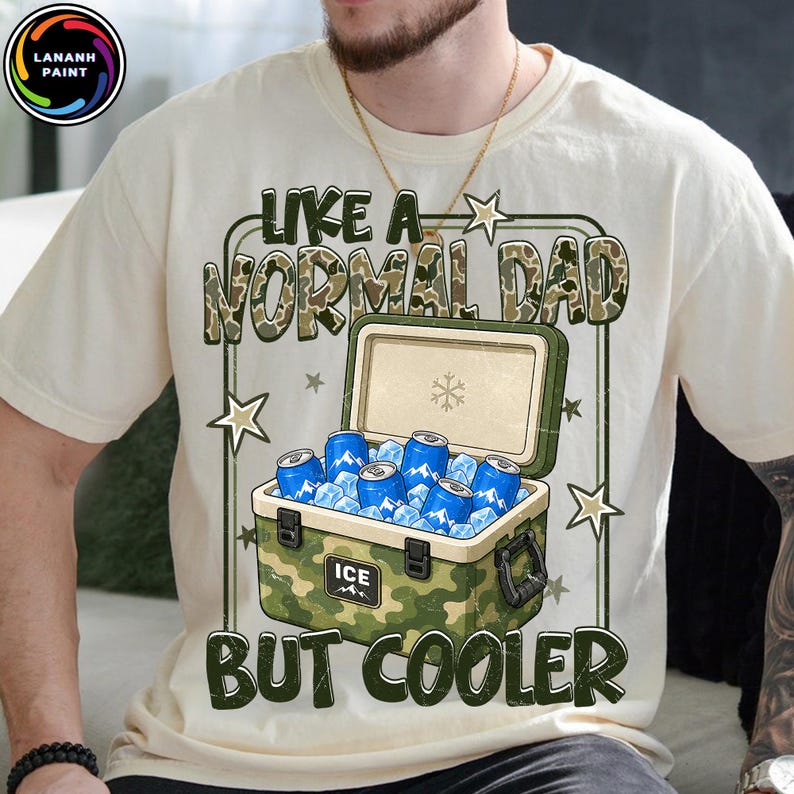 K&ouml;nnte beinhalten: Cremefarbenes T-Shirt mit dem Text "LIKE A NORMAL DAD BUT COOLER" &uuml;ber einer K&uuml;hlbox, die mit blauen Dosen und Eis gef&uuml;llt ist. Die K&uuml;hlbox hat ein Tarnmuster. Das Shirt hat einen Rundhalsausschnitt und kurze &Auml;rmel.
