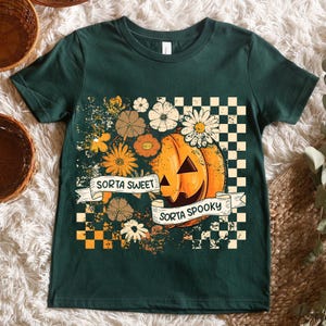 May include: Dark green t-shirt featuring a Halloween-themed design. The graphic includes a jack-o'-lantern, flowers, and the phrases "Sorta Sweet" and "Sorta Spooky" on banners. A checkered pattern frames the design.