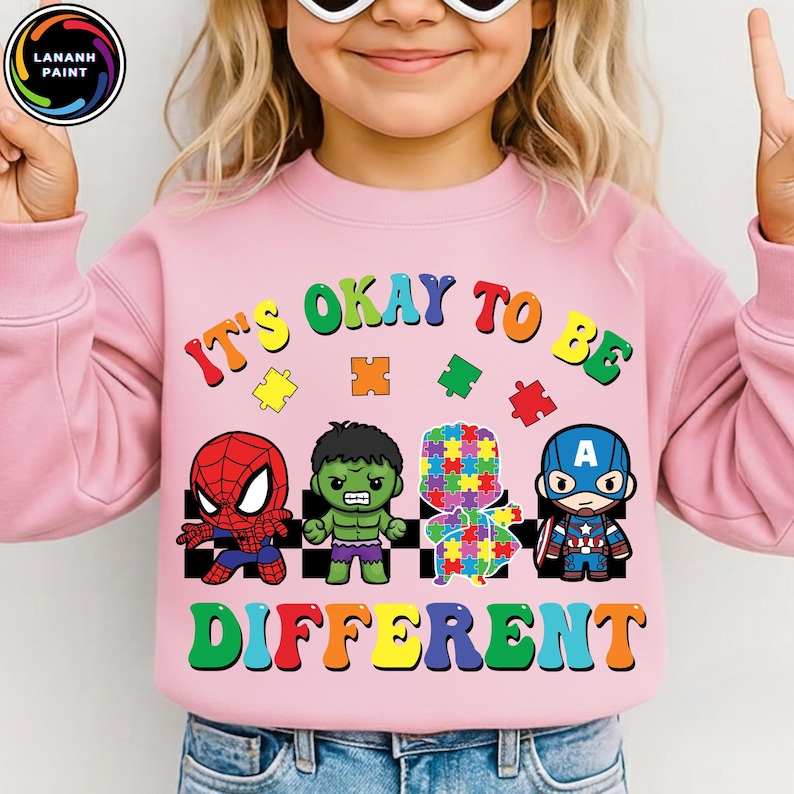 May include: Pink sweatshirt with the text "It's Okay to Be Different" and cartoon images of Spider-Man, Hulk, a puzzle piece figure, and Captain America. The sweatshirt has a crew neck and long sleeves.
