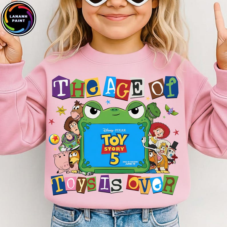 Puede incluir: Una sudadera rosa claro con un estampado gr&aacute;fico que incluye el texto "THE AGE OF TOYS IS OVER" y "TOY STORY 5". El dise&ntilde;o presenta un marco verde con forma de rana que rodea el t&iacute;tulo de la pel&iacute;cula, junto a varios personajes.