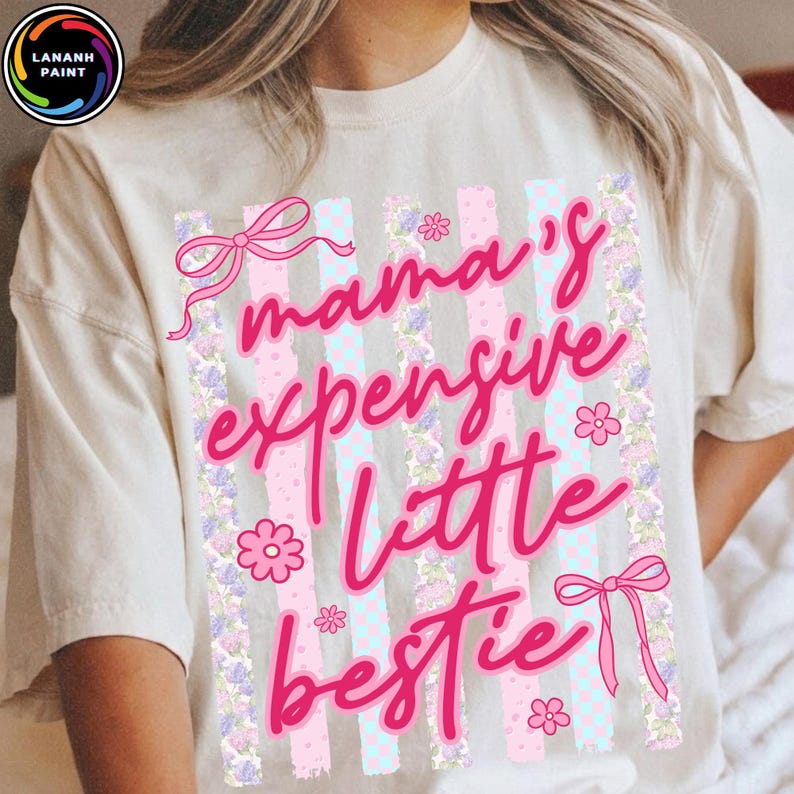 Puede incluir: Camiseta color crema con el texto rosa "mama's expensive little bestie". El dise&ntilde;o incluye rayas rosas y azules, motivos florales y lazos.