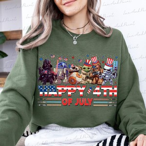 K&ouml;nnte beinhalten: Gr&uuml;nes Sweatshirt mit einer Grafik, die Star Wars-Charaktere Darth Vader, R2-D2, Grogu, Chewbacca und einen Sturmtruppler zeigt, die alle patriotische H&uuml;te tragen. Der Text "HAPPY 4TH OF JULY" ist in Rot, Wei&szlig; und Blau dargestellt.