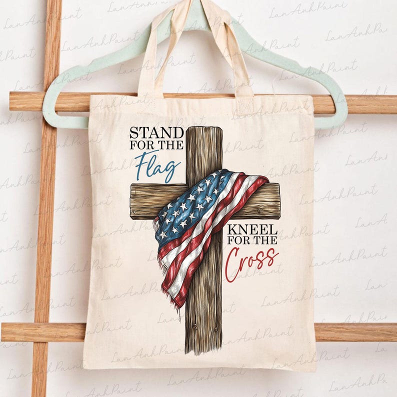 Puede incluir: Una bolsa de mano beige con un dise&ntilde;o de cruz de madera. La cruz est&aacute; cubierta con una bandera estadounidense. El texto en la bolsa dice "STAND FOR THE Flag" y "KNEEL FOR THE Cross". La bolsa est&aacute; colgada en un estante de madera.