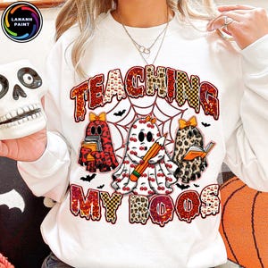 May include: White sweatshirt with a Halloween-themed graphic. The design features three ghost characters, a spider web, bats, and the text "TEACHING MY BOOS". The ghosts have patterns like cherries, leopard print, and plaid.